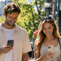 Couple walks down a sunny street with shopping bags while checking their phones.