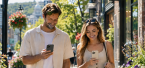 Couple walks down a sunny street with shopping bags while checking their phones.